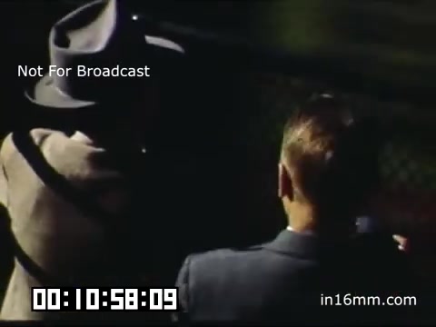 The still from the 16mm Kodachrome footage of a Tigers game at Briggs Stadium captures a moment with two men in suits, one wearing a hat. They are seen from behind, likely watching a game, with the stadium's backdrop visible. The colors are rich, typical of Kodachrome film from that era, giving a nostalgic quality to the scene. The timecode indicates a specific moment within the footage, and the caption notes that it is not for broadcast.