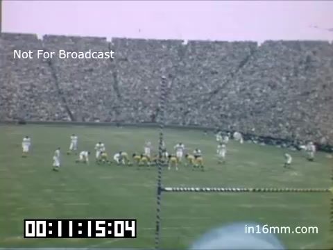 The still from 'University of Michigan Wolverines 1948-1950' shows a football game setting, featuring players in traditional uniforms on the field. The vast stadium is filled with an audience, highlighting the energetic atmosphere of the event. The image captures a moment just before a play, with a scoreboard displaying the time. The scene reflects a classic college football experience, marked by its historical significance.