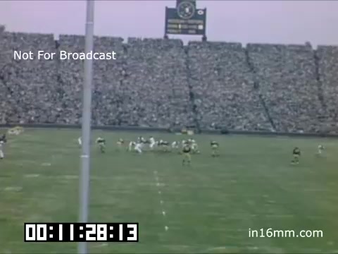 The still captures a football game featuring the University of Michigan Wolverines from the late 1940s to 1950. A packed stadium is visible, with fans filling the stands. In the foreground, players from both teams are positioned on the field, with action likely underway. An electronic scoreboard is prominently displayed in the background. The scene is presented in a vintage color style typical of 16mm film from that era. The time code indicates the moment in the film.