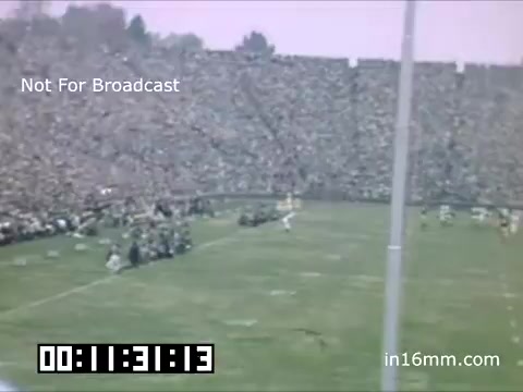 The still features a vibrant scene from a University of Michigan Wolverines football game between 1948 and 1950. The image captures a large crowd in the stadium, showcasing a lively atmosphere. Players are positioned on the field, and the action suggests a moment of excitement during the game. The color and graininess are characteristic of 16mm film from that era, reflecting the historical context.