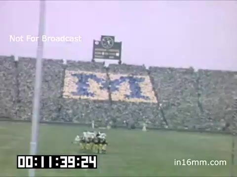 The still features a packed stadium during a University of Michigan Wolverines football game from 1948-1950. The crowd has arranged themselves to form a large 'M' in blue and maize, symbolizing school spirit. The scoreboard is visible in the background, contributing to the game-day atmosphere. The scene captures the excitement and camaraderie of college football, with players positioned on the field. The date and time in the corner indicate the moment captured.