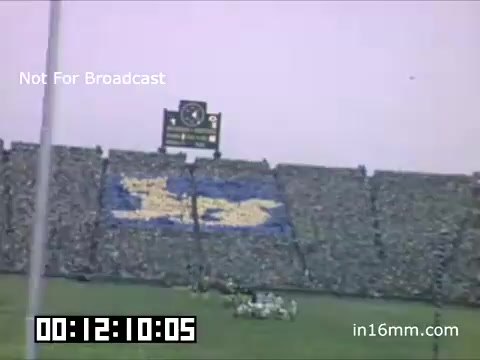 The still captures a scene from a University of Michigan Wolverines football game between 1948 and 1950. A large crowd fills the stands, showcasing a vibrant blue and maize color scheme. In the background, a scoreboard displays the game's status. The atmosphere is energetic, indicative of a lively sports event.