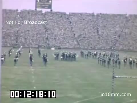 The still depicts a lively scene from a University of Michigan Wolverines event between 1948 and 1950. The foreground shows a marching band or drill team performing on a football field, while a large crowd fills the stands in the background. The atmosphere is vibrant, likely during a halftime show or parade, highlighting school spirit and tradition. The image has a vintage quality typical of 16mm film, capturing a moment in collegiate history.