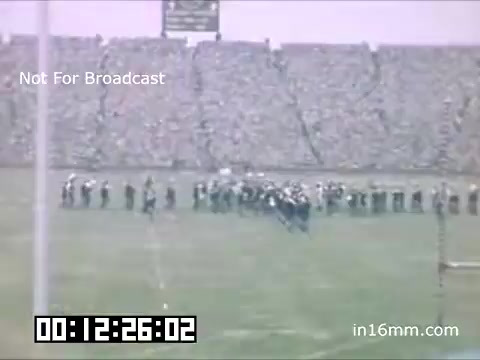 The still captures a scene from a 16mm film featuring the University of Michigan Wolverines between 1948 and 1950. It depicts a football field populated with members of a marching band or pep group, suggesting a spirited event, likely a pre-game or halftime performance. The grandstands are filled with spectators, emphasizing the crowd's engagement. The bottom of the frame displays a timestamp and a 'Not For Broadcast' label, indicating the film is for archival or private viewing only.