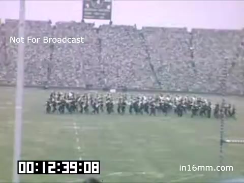The still depicts a large crowd at a sports event, likely a football game, featuring the University of Michigan Wolverines from the late 1940s to early 1950s. In the foreground, a band or group of performers is organized in formation on the field. The vibrant atmosphere suggests a festive occasion, possibly a halftime show, with the audience enthusiastically engaged. The timestamp indicates it's part of a longer film sequence. The image is marked as 'Not For Broadcast,' emphasizing its archival nature.