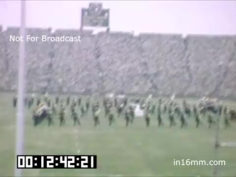 The still captures a scene from a University of Michigan Wolverines event between 1948 and 1950, featuring a large crowd in a stadium. A marching band or performance group is visible on the field, showcasing a coordinated routine. The atmosphere appears festive, with the audience engaged in the background. The color tone suggests a nostalgic, vintage quality, typical of 16mm film from that era.