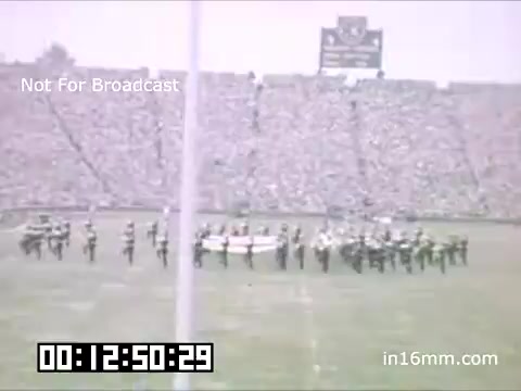 The still captures a lively scene from a University of Michigan Wolverines event between 1948 and 1950, showcasing a marching band performance on the football field. The band members are arranged in formation, likely part of a halftime show, with a packed stadium of spectators in the background. The atmosphere reflects the enthusiasm and spirit typical of college football games. The timestamp indicates the specific moment within the film.