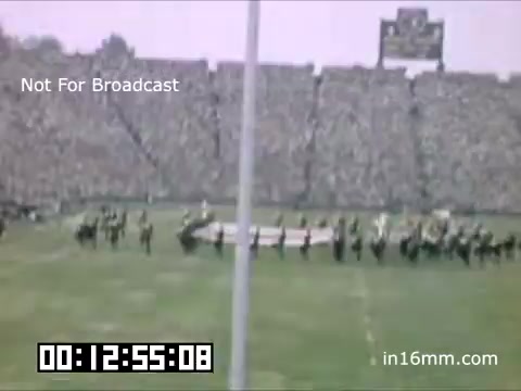 The still captures a scene from a University of Michigan Wolverines event between 1948 and 1950. It features a performance on the football field, likely during halftime. A marching band or performance group is arranged on the grass, with a substantial crowd visible in the stadium stands, creating an atmosphere of excitement. A scoreboard is faintly seen in the background, indicating the venue's sports context.