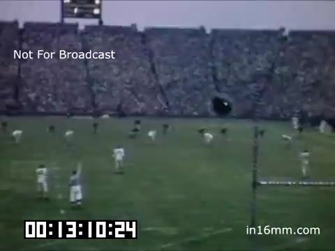 The 16mm still depicts a football game featuring the University of Michigan Wolverines, likely from the late 1940s to early 1950s. The scene captures players on the field, with a packed stadium in the background, indicative of a lively atmosphere. The footage is in color, showcasing vintage sports aesthetics, with the timestamp and 'Not For Broadcast' watermark indicating it is for archival purposes.