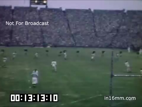 The still depicts a crowded football stadium during a game, likely featuring the University of Michigan Wolverines from the 1948-1950 era. Players are seen on the field in vintage uniforms, with a vibrant audience filling the stands in the background. The image carries a nostalgic vibe, reflecting the energy of a mid-20th century sporting event. The timestamp suggests it's part of a longer film.