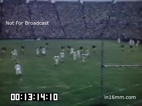 The still depicts a scene from a 1948-1950 University of Michigan Wolverines football event, showcasing players on the field in action. The stadium is filled with spectators, creating a vibrant atmosphere. A scoreboard or timer is visible, indicating the time elapsed in the footage. The overall mood captures the excitement of a college football game during this era, complemented by the accompanying music from Kevin MacLeod.