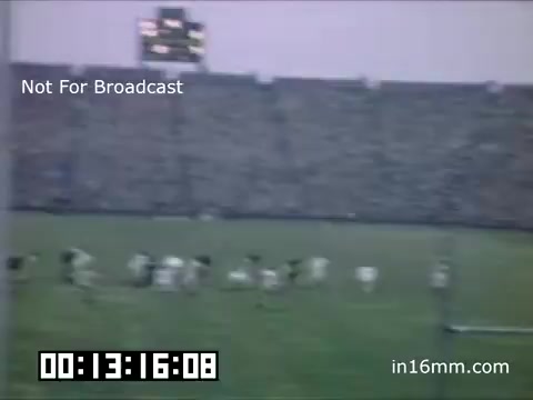 The still features a scene from a football game involving the University of Michigan Wolverines, captured between 1948 and 1950. The image shows players on the field, with a large crowd in the stadium stands. A scoreboard is visible in the background, indicating the game's progress. The overall ambiance suggests a lively sports event, typical of college football traditions during that era.