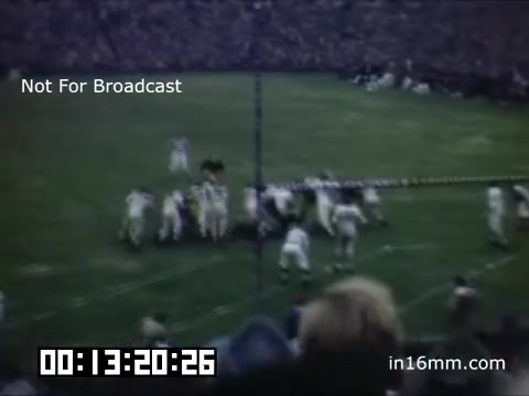 The still from 'University of Michigan Wolverines 1948-1950' captures a football game scene, likely illustrating an intense moment of action on the field. Players in white uniforms can be seen in a tactical formation, while spectators fill the stands surrounding the field. The image has a vintage quality, characteristic of 16mm film, and the overlay text indicates it is not meant for broadcast. The timestamp at the bottom suggests it is part of a larger film sequence.