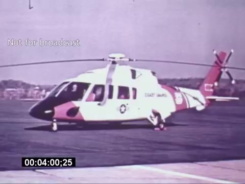 The still shows a Sikorsky S-76 helicopter parked on a tarmac, emphasizing its sleek design and distinctive coloring. The helicopter features a white body with red and black accents, and bears markings indicating its use by the Coast Guard. The background hints at a clear day, possibly at an airfield or base. The image is labeled 'Not for broadcast,' suggesting it was intended for promotional or archival purposes, and the timestamp indicates a moment during the film.