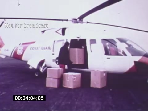 The still from the 'Sikorsky S-76 Promotional Film' shows a Sikorsky S-76 helicopter with the markings of the Coast Guard. In the image, a person is seen unloading several boxes from the open door of the helicopter, which is parked on a flat surface. The setting appears to be a cloudy day, providing a muted backdrop for the scene. The visual suggests a focus on the helicopter's utility and operational capabilities, particularly in cargo transport or rescue scenarios.