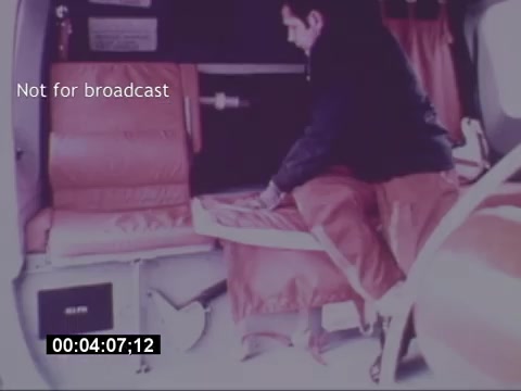 The still from the 'Sikorski S-76 Promotional Film' features a person inside the interior of a Sikorsky S-76 helicopter. The individual is seen adjusting or folding a seat, which is upholstered in a vibrant orange material. The cabin appears spacious and designed for comfort. A timestamp indicates the scene is approximately four minutes and seven seconds into the film. The text 'Not for broadcast' suggests that this material is intended for private or promotional use rather than public airing.