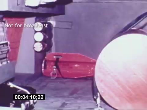 The still from the 'Sikorski S-76 Promotional Film' depicts a close-up view of an interior setting, possibly within a helicopter. The image features various industrial components, including what appears to be a large, cylindrical object and a red item that resembles a life vest or flotation device. There are also several circular elements that suggest instrumentation or controls. The color palette is primarily muted, with shades of gray and red, giving a technical and functional feel to the scene. The overlay text 'Not for broadcast' indicates that this image is intended for private viewing only.