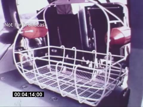 The still from the 'Sikorski S-76 Promotional Film' features a close-up view of a helicopter interior, specifically showcasing an empty wire basket possibly used for cargo or equipment storage. Red emergency flotation devices are attached to the sides, indicating the safety features of the aircraft. The background appears to include cockpit components or controls, hinting at the operational context of the helicopter. The frame is timestamped and has a 'Not for broadcast' watermark, suggesting it's for archival or promotional purposes rather than general release.