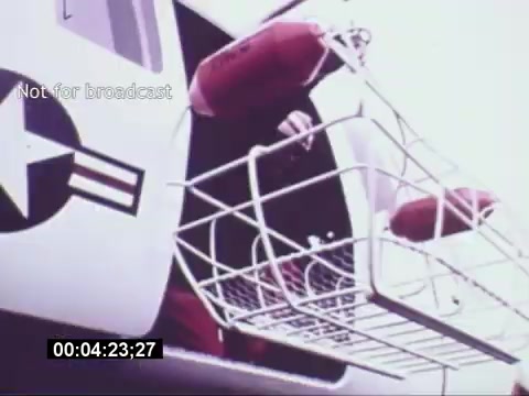 The still from the 'Sikorski S-76 Promotional Film' shows a Sikorsky S-76 helicopter with a focus on its open door. A rescue basket hangs from the door, indicating its use for medical or emergency services. The background features a military insignia, suggesting a connection to military operations. The overall tone appears promotional, emphasizing the helicopter's capabilities. The absence of a soundtrack adds a layer of silence to the visual presentation.