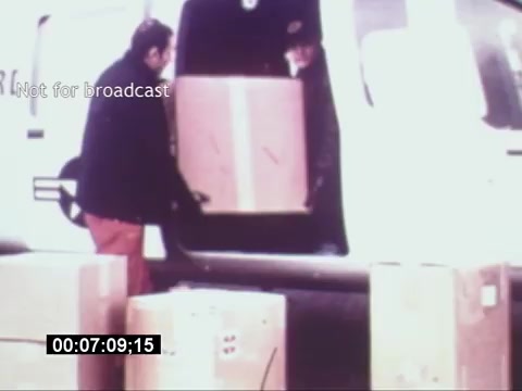 The still from the 'Sikorski S-76 Promotional Film' depicts two individuals loading large boxes into a helicopter. One person stands in the open doorway of the aircraft, while the other is seen outside, lifting a box. The environment appears to be a logistics or transport setting, showcasing the Sikorsky S-76's utility in cargo operations. The image lacks sound, and the timestamp suggests it is a specific moment in the sequence, emphasizing the efficiency of the loading process.