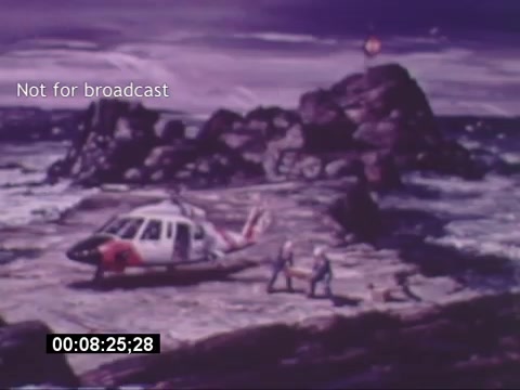 The still from the 'Sikorski S-76 Promotional Film' features a Sikorsky S-76 helicopter on a rocky coastline. Three figures are seen, likely personnel or crew, transporting a stretcher across the rugged terrain. The scene suggests a rescue or emergency operation, highlighting the helicopter's utility in challenging environments. The backdrop is dramatic, with a turbulent sea and overcast sky, enhancing the sense of urgency and purpose. The image is marked 'Not for broadcast,' indicating it is for promotional or internal use only.