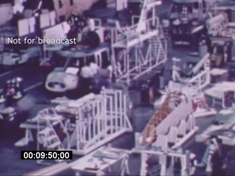 The still from the Sikorski S-76 promotional film depicts a busy manufacturing or assembly area. Various components and structures related to helicopter production are visible, along with a vehicle, likely used for transportation within the facility. The image is presented in a vintage color palette, typical of 16mm film, suggesting a focus on the industrial process and craftsmanship involved in creating the Sikorski S-76. A timestamp and the note 'Not for broadcast' indicate it's meant for specific viewing contexts rather than general release.