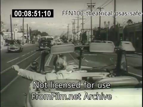 The still shows a busy street scene from a 1949 theatrical public service announcement promoting safe driving. A person is seen in a convertible, signaling with their arm, while various cars and power lines populate the background. The era's vintage vehicles and the overall ambiance reflect mid-20th-century America, emphasizing the importance of road safety awareness.