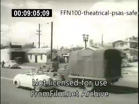 The still from the 1949 theatrical public service announcements for safe driving features a scene with a white convertible car and a large truck on a road. The backdrop includes houses and power lines, suggesting a suburban environment. The image emphasizes the importance of road safety during that era, likely showcasing a message related to cautious driving practices. The overall aesthetic reflects the style and atmosphere of late 1940s America.