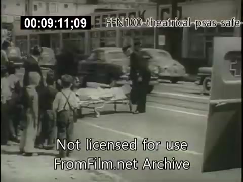 The still depicts a scene from a 1949 public service announcement emphasizing safe driving. It shows a group of children watching as a person is being assisted on a stretcher, implying the consequences of unsafe driving. The backdrop features vintage cars and urban architecture reflective of the era, reinforcing the seriousness of road safety awareness.