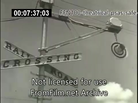 The still depicts a railroad crossing sign prominently, with an arm signaling the crossing. The sky is clear, giving a sense of a typical day. This image aligns with the 1949 theatrical public service announcements promoting safe driving, emphasizing caution around railroad crossings.