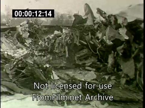 This still captures the aftermath of the N6907C Lockheed Constellation crash on December 16, 1960. The image depicts a wreckage site, showing fragments of the aircraft scattered and in disarray. Debris is strewn across a bleak landscape, indicating the severity of the crash caused by a mid-air collision with a DC aircraft. The black-and-white footage reflects the tragic and chaotic scene, characteristic of news footage from that era.