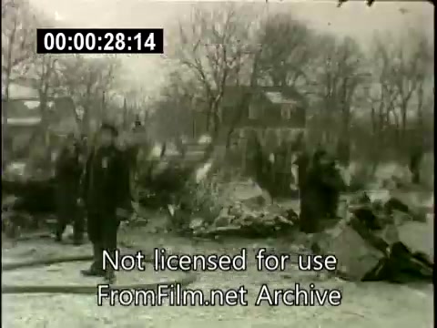 The still captures a scene from the aftermath of the Staten Island plane crash on December 16, 1960. The image shows emergency personnel and onlookers surveying the wreckage of the crash site, with debris scattered on the ground. In the background, snow-covered trees and residential buildings appear, indicating a suburban environment. The atmosphere is somber, reflecting the gravity of the tragedy. The footage was distributed by United Press to TV stations, marking a significant historical event in aviation safety.