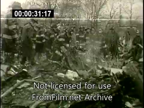The still depicts a grim scene from the aftermath of the Staten Island Plane Crash on December 16, 1960. In the image, a group of individuals, likely emergency responders and onlookers, gathers around the wreckage of the Lockheed Constellation aircraft. The area is marked by debris scattered across the ground, with trees in the background. The somber atmosphere reflects the tragic nature of the event, which resulted from a mid-air collision. The footage is attributed to United Press and likely intended for dissemination to TV stations for coverage.