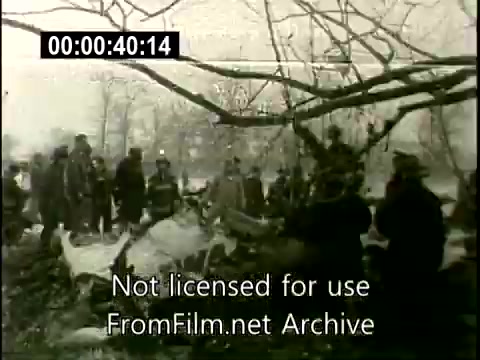 The still shows the aftermath of the Lockheed Constellation crash on December 16, 1960, featuring a scene of chaos and devastation. In the foreground, debris from the aircraft is visible, partially covered by branches. A group of onlookers and emergency personnel gather around the crash site, their figures silhouetted against a bleak, overcast sky. The atmosphere appears somber, reflecting the tragic nature of the incident.