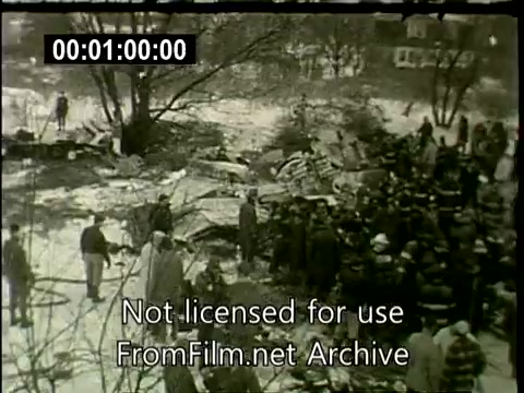 The still depicts the aftermath of the N6907C Lockheed Constellation crash in Staten Island on December 16, 1960. It shows a chaotic scene with emergency responders and onlookers gathered around the wreckage in a snow-covered area. Fragments of the aircraft are scattered, and trees are visible in the background. The presence of officials in uniforms indicates an active rescue and investigation effort. Overall, the image captures a somber moment following a tragic aviation incident.