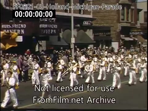The still captures a vibrant scene from the Holland, Michigan Tulip Parade, circa 1948-1950. A large group of parade participants in white uniforms is seen marching down the street, showcasing a festive atmosphere. Local businesses line the background, with spectators gathered to enjoy the celebration. The bright colors typical of Kodachrome film enhance the lively ambiance of this community event.
