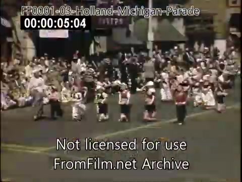 The still captures a vibrant scene from the Holland, Michigan Tulip Parade, circa 1948-1950. Participants in colorful costumes march along a street lined with spectators. The atmosphere is festive, with a mix of marching bands and dancers, showcasing the community's celebration of Dutch heritage through tulip-themed festivities. Historical storefronts and a diverse crowd further enhance the lively setting of this iconic event.
