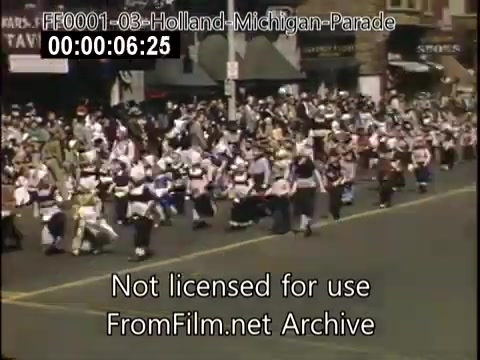 The still captures a lively scene from the Holland, Michigan Tulip Parade, dating back to circa 1948-1950. It features a colorful procession of participants in traditional attire, likely celebrating Dutch heritage, as they march along a crowded street. Spectators line the sidewalks, creating a festive atmosphere filled with excitement and community spirit. The vibrant colors and lively movement evoke the charm of mid-20th-century parades.