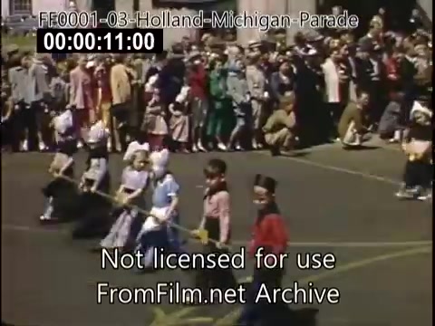 The still captures a vibrant moment from the Holland, Michigan Tulip Parade, likely between 1948 and 1950. In the foreground, children dressed in traditional costumes are participating in the parade, while a large crowd of spectators eagerly watches from the sidelines, dressed in mid-20th-century attire. The scene is lively, showcasing the cultural celebration and community spirit of the event.