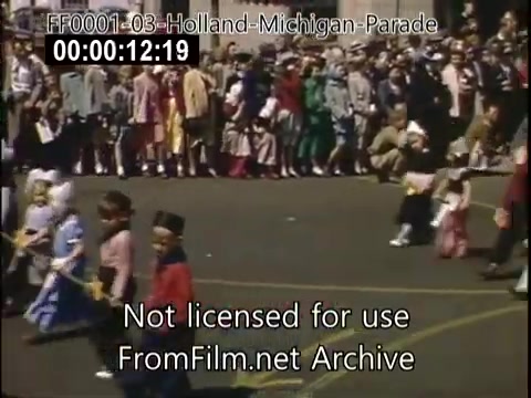 The still captures a vibrant scene from the Holland, Michigan Tulip Parade, likely dating between 1948 and 1950. A diverse crowd lines the street, watching a procession of children dressed in colorful costumes. The lively atmosphere reflects the celebration of Dutch heritage, with many participants showcasing traditional attire. The use of Kodachrome film enhances the vivid colors, adding to the festive mood of the event.