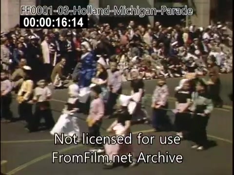 The still captures a vibrant scene from the Holland, Michigan Tulip Parade circa 1948-1950. In it, a diverse group of participants dressed in colorful traditional attire march along a street lined with a large crowd of spectators. The festive atmosphere is underscored by the presence of tulips and decorative elements, reflecting the cultural celebration of the event. The image is characterized by rich Kodachrome colors, showcasing the lively spirit of the parade.