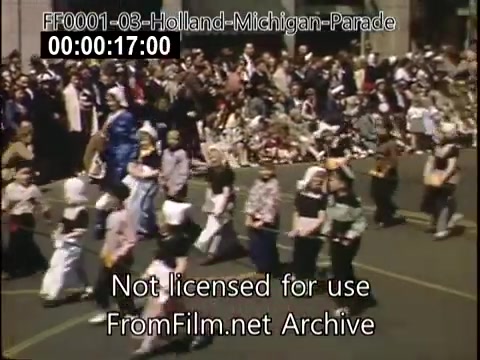 The still captures a vibrant scene from the Holland, Michigan Tulip Parade circa 1948-1950. A procession of children and adults in colorful traditional costumes can be seen marching down the street. The crowd in the background is lively, with many spectators enjoying the festivities. The image features a mix of expressions and clothing styles, indicative of the era, highlighting the community spirit of the celebration.
