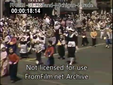 The still from the 'Holland, Michigan Tulip Parade' captures a vibrant scene from the late 1940s to early 1950s. Participants, likely in traditional Dutch attire, are seen marching in a festive parade setting. In the background, a large crowd is gathered along the parade route, enjoying the celebration. The colorful clothing and lively atmosphere reflect the cultural significance of the event, which celebrates Dutch heritage through music, dance, and flowers.