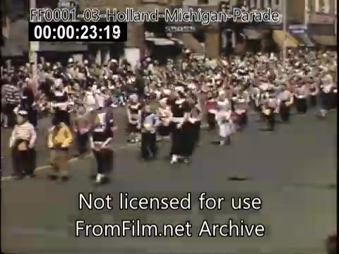 The still captures a lively scene from the Holland, Michigan Tulip Parade, taking place between 1948 and 1950. A large crowd lines the streets, with participants dressed in traditional attire, including costumes reflective of Dutch heritage. Colorful banners and decorations enhance the festive atmosphere, showcasing a vibrant community celebration. The image highlights the cultural significance of the event, drawing both locals and visitors.