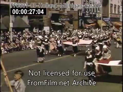 The still captures a vibrant scene from the Holland, Michigan Tulip Parade, circa 1948-1950. Participants in traditional attire, likely honoring Dutch heritage, are seen marching with large flags. A lively crowd lines the street, showcasing enthusiastic spectators enjoying the festive atmosphere. The colorful display reflects the celebration's cultural significance, with storefronts adding to the historic backdrop.