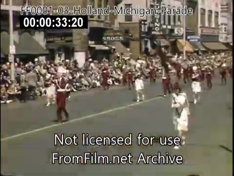 The still captures a vibrant scene from the Holland, Michigan Tulip Parade, likely between 1948 and 1950. It features a procession with performers dressed in colorful costumes, including red and white outfits. A large crowd is gathered along the street, watching the parade amidst a backdrop of storefronts. The atmosphere is festive, highlighting the community's celebration of Dutch heritage and the blooming of tulips.