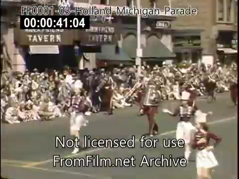 The still captures a lively scene from the Holland, Michigan Tulip Parade in the late 1940s. A crowd gathers along the street, with spectators enjoying the festivities. Participants in the parade include groups of marchers, dressed in vibrant attire, and possibly a marching band, contributing to the festive atmosphere. The background features storefronts, adding a quaint charm typical of that era. The scene is a colorful representation of community celebration with a focus on cultural heritage.