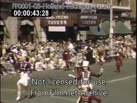 The still depicts a vibrant scene from the Holland, Michigan Tulip Parade, circa 1948-1950. Participants, possibly in festive costumes, are seen marching along a street lined with spectators. The background features buildings, including a tavern, indicating a lively community atmosphere. The overall scene exudes a celebratory spirit typical of a parade, enhanced by the colorful attire and the gathering of onlookers.