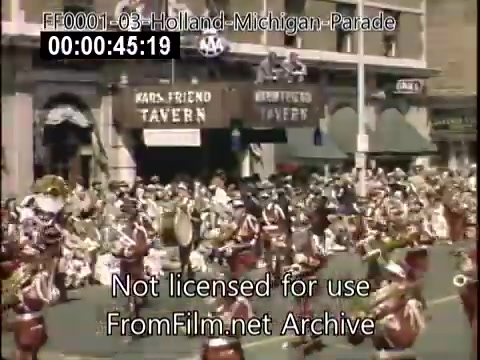 The still depicts a vibrant scene from the Holland, Michigan Tulip Parade, likely between 1948 and 1950. A large crowd is gathered on a city street, with participants in traditional attire, including musicians and dancers. The backdrop features the 'Kars-Friend Tavern' and other buildings, emblematic of the era. The festive atmosphere highlights the celebration of Dutch heritage, as evidenced by the colorful costumes and floral decorations.