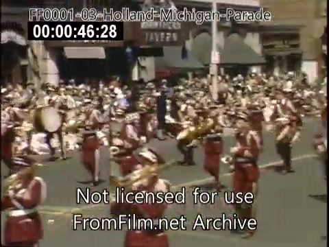 The still depicts a vibrant scene from the Holland, Michigan Tulip Parade, likely between 1948 and 1950. A marching band in uniforms of red and white is prominently featured, while a dense crowd of spectators lines the street. Buildings and storefronts in the background hint at the small-town atmosphere of the parade, enhancing the festive and communal spirit of the event. The color is vivid, typical of Kodachrome film, showcasing the lively energy of this mid-century celebration.